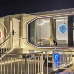 A close-up of a lit-up prefab house at night, with a curved balcony and illuminated outdoor elements.