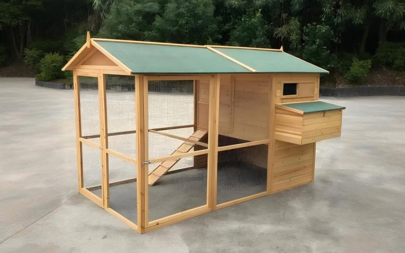 A wooden chicken coop with a green roof, wire fencing, and a small ladder inside, set on a concrete surface.