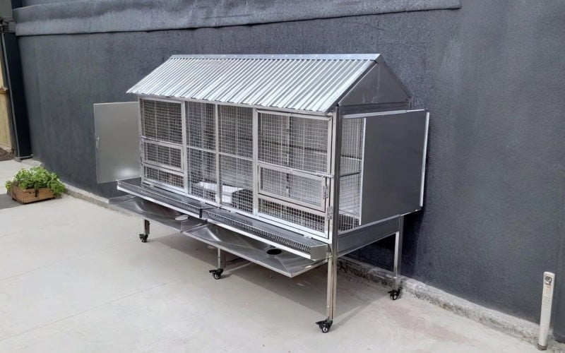 A metal chicken coop with a sloped roof, wire mesh walls, and wheels, placed against a gray wall.