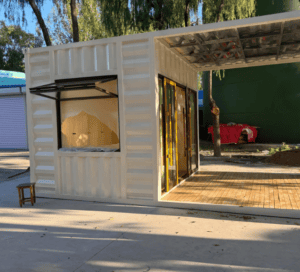 A compact white shipping container home with a sliding glass door, an awning-style window, and a covered wooden patio, set among trees.