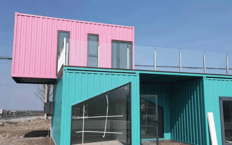 Colorful modular shipping container house with pink and teal units, featuring large glass windows and a rooftop balcony.