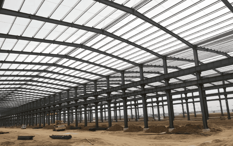 A wide steel warehouse under construction, featuring a high-ceiling structure with steel beams and a transparent roof.