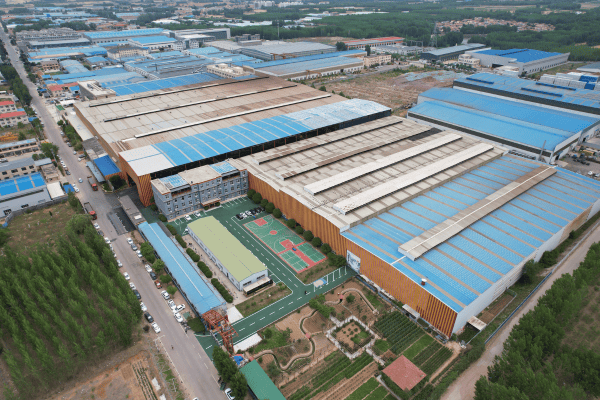 An aerial view of a large factory complex with multiple blue-roofed buildings, green landscaping, and parking areas.