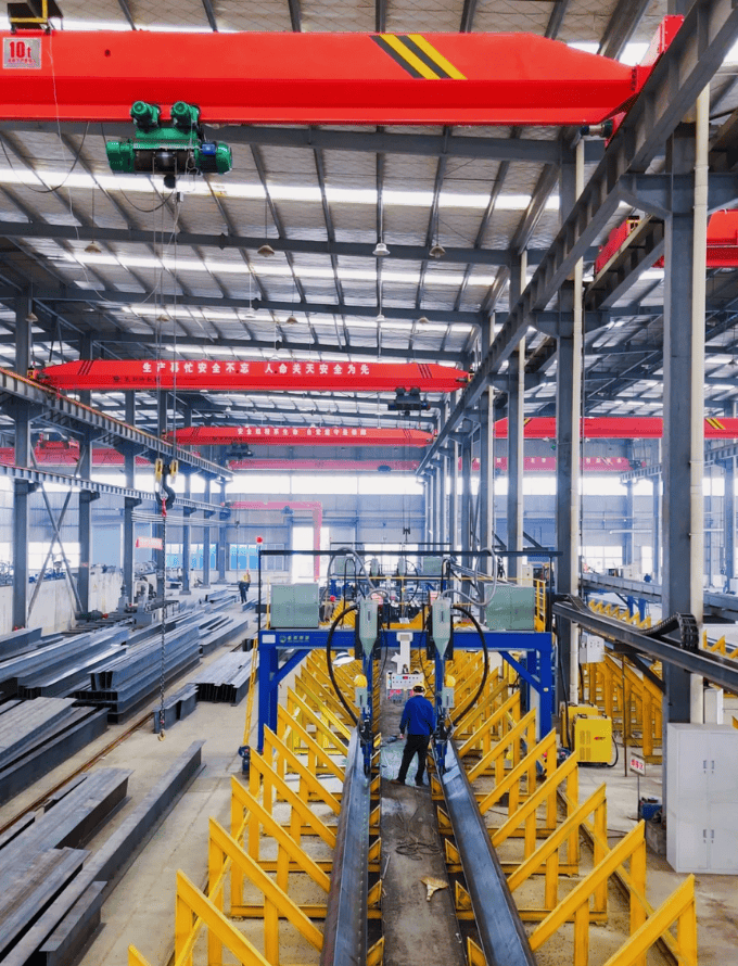 Interior of a steel structure workshop featuring red overhead cranes, steel beams, and workers operating machinery for steel fabrication.