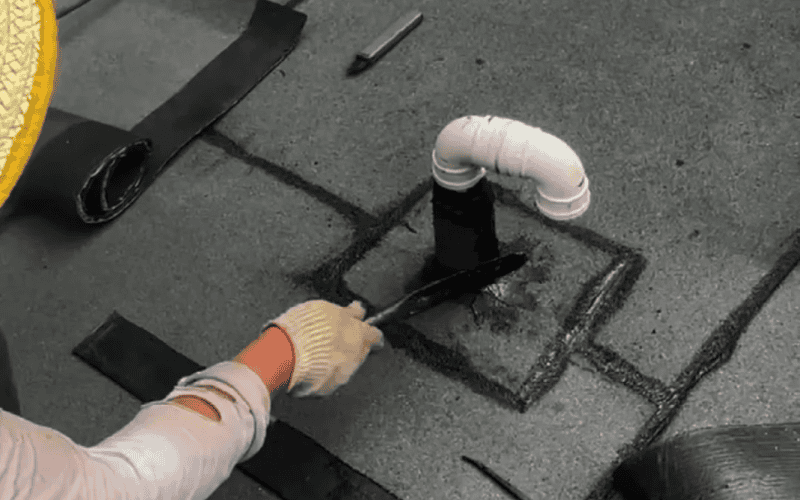 A worker in gloves applying sealant around a ventilation pipe on a textured roof surface to prevent leaks.