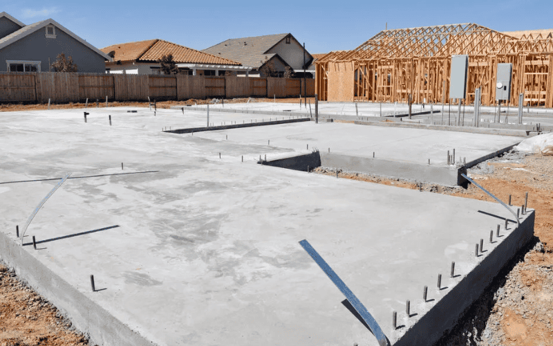 A residential concrete slab foundation with embedded bolts, surrounded by partially constructed wooden frames and nearby houses.