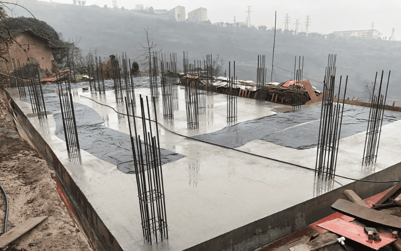 A wet concrete foundation with exposed vertical steel reinforcements, set against a hilly background and foggy weather.