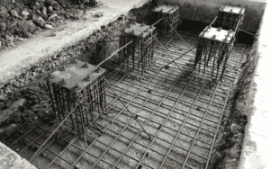 A reinforced concrete foundation under construction, featuring a rebar grid and steel column base plates with vertical reinforcements, prepared for structural support.