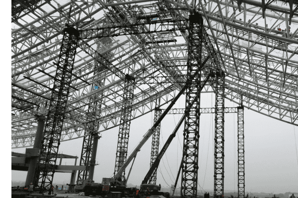 A large steel structure under construction, supported by cranes and scaffolding, showcasing intricate framework details.