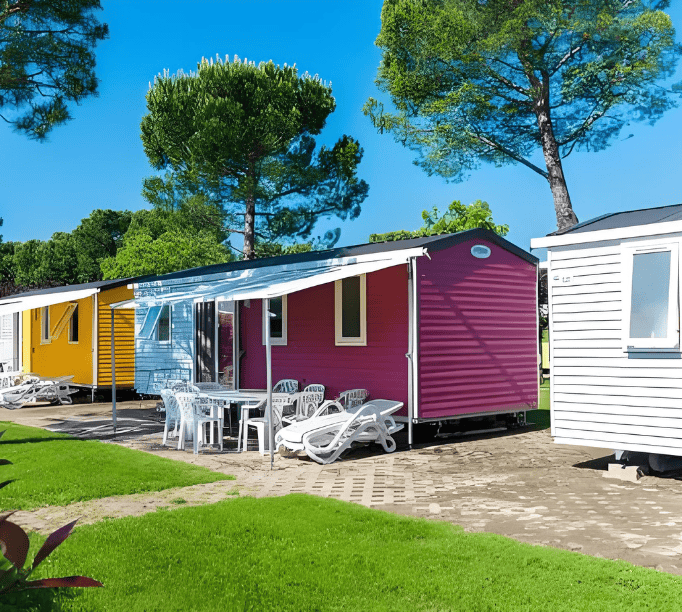 A colorful row of mobile homes, including pink, yellow, and white houses, surrounded by lush green trees and grassy areas, with seating and a small table in front of one house.