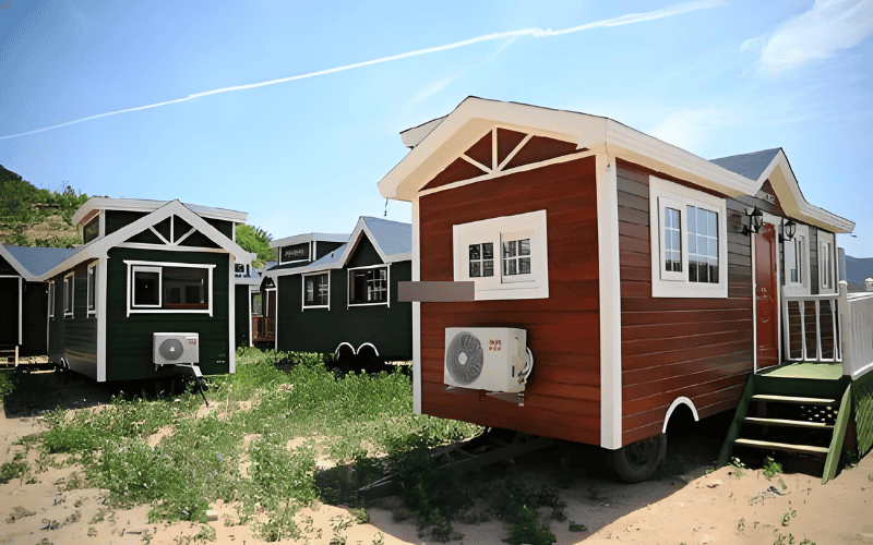A group of colorful mobile homes with a mix of green, red, and white exteriors, featuring windows and a front porch area surrounded by plants and open land.