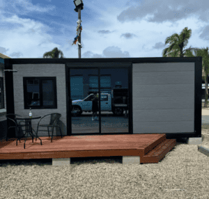 Modern modular home with dark trim and wood deck, featuring a small outdoor table and chairs, set against a background of palm trees and blue sky.