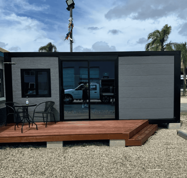 Modern modular home with dark trim and wood deck, featuring a small outdoor table and chairs, set against a background of palm trees and blue sky.