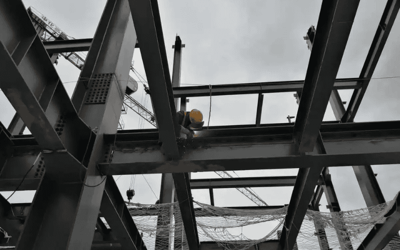 A construction worker welding a steel framework for a large pole barn while secured with safety gear at an elevated position.