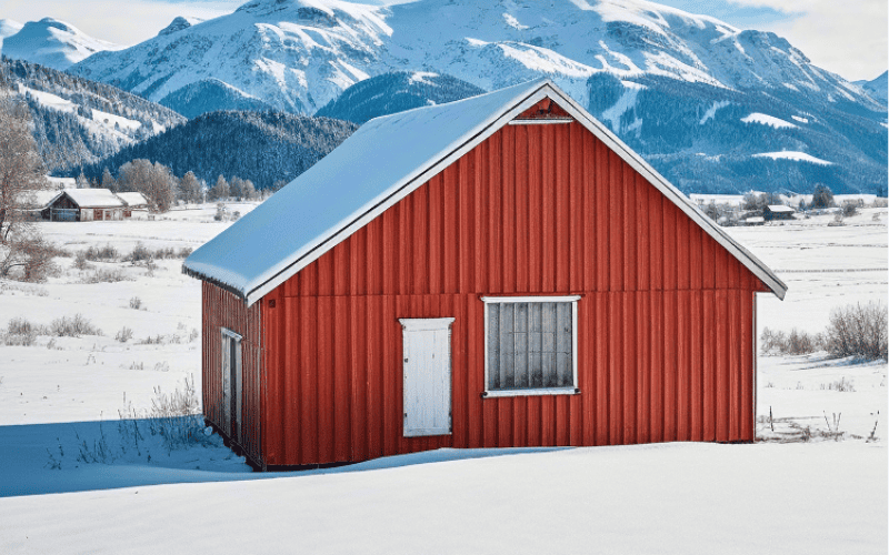 A red steel salt barn in a snowy rural landscape with a white door and window. Snow covers the ground, with mountains and farmhouses in the background.