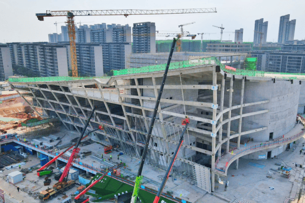 A large steel structure building under construction, with cranes and Urban high-rises int and background.