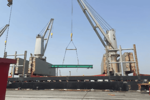 A large green steel beam being hoisted by cranes at a shipping port under a bright blue sky.