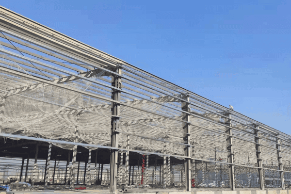 A steel structure under construction with visible beams and netting, set against a clear blue sky.