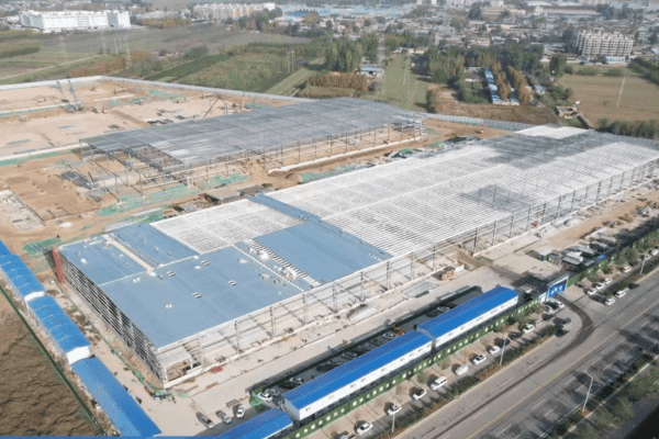 An aerial view of a large logistics facility under construction, featuring multiple steel frames and a completed section with a blue roof.