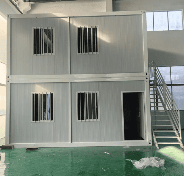 This image shows a two-story container office with metal windows, a door, and a staircase leading to the upper level, set in a spacious industrial interior. The floor is green, and construction materials are scattered around.