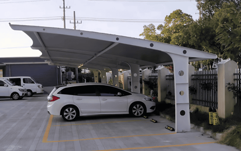 A white metal lean-to carport sheltering vehicles, supported by curved steel beams with circular cutouts.