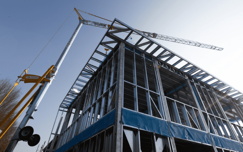 Light steel framing of a building under construction with a crane lifting components against a clear sky.