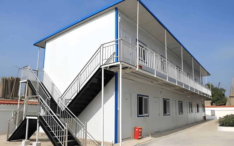 A two-story prefabricated modular building with white walls, blue trim, and an external staircase, situated in an open courtyard under a clear sky.