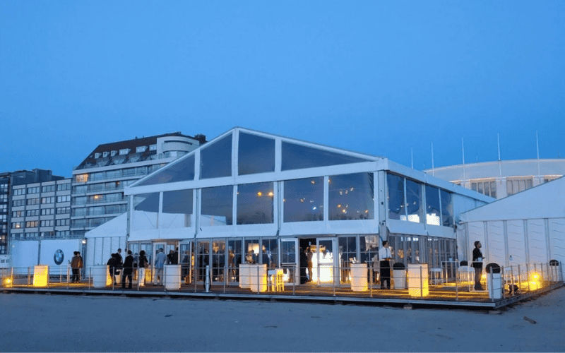 An event tent with clear windows, lit up in the evening, with people gathered outside.