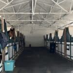 Interior of a metal horse barn with horses in stalls, showing brick - and - metal partitions, equipment hanging, and a wide central aisle.