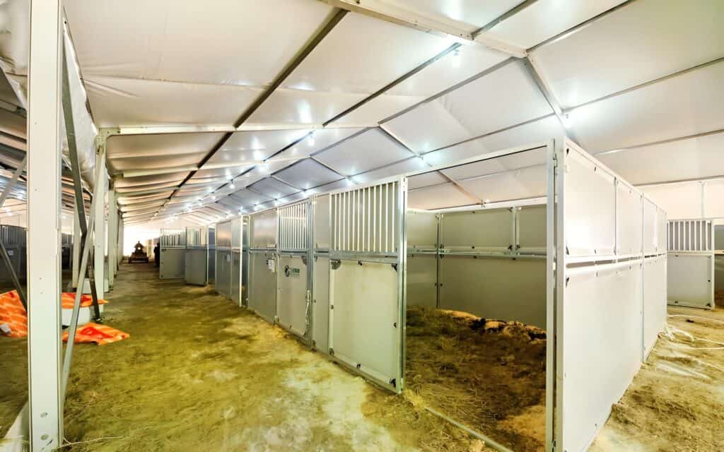 Another view of a metal horse barn kit installation, featuring empty stalls, a sandy floor, and bright overhead lighting.