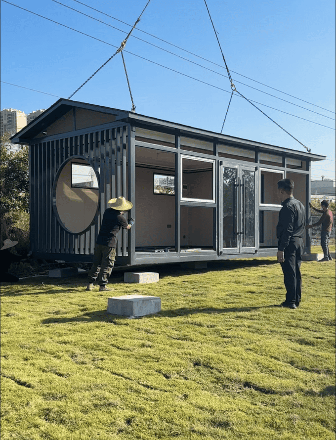 A modular home being lifted into place by a crane, with workers guiding the process. The house features large windows and a circular design element, set against a clear blue sky.