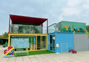 Multi-level modular daycare made from repurposed shipping containers, featuring a rooftop play area, outdoor decorations, and bright, child-friendly design.