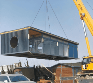 A mobile home being moved on a flatbed truck with a crane assisting in positioning, highlighting the mobility aspect of manufactured housing.