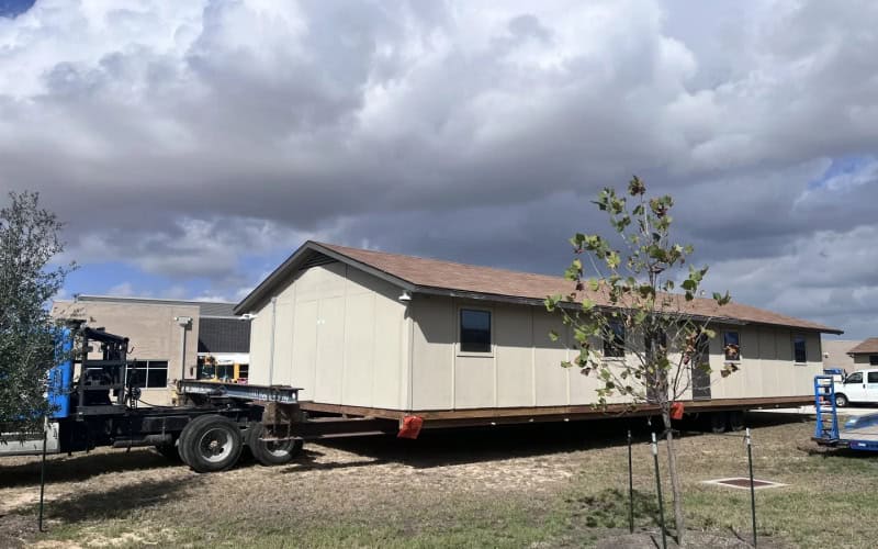 A beige portable classroom on a flat - bed truck, ready for transport, with trees and a building in the background under a cloudy sky.