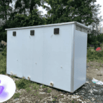 White portable restroom unit with a simple design, located outdoors among greenery with visible vents.