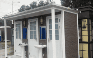 A portable restroom unit with three separate stalls, each featuring a washbasin and blue accent windows, set outdoors.