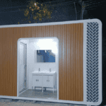 A prefab ablution block with a modern design, featuring a wooden accent wall and twin washbasins, illuminated at night.