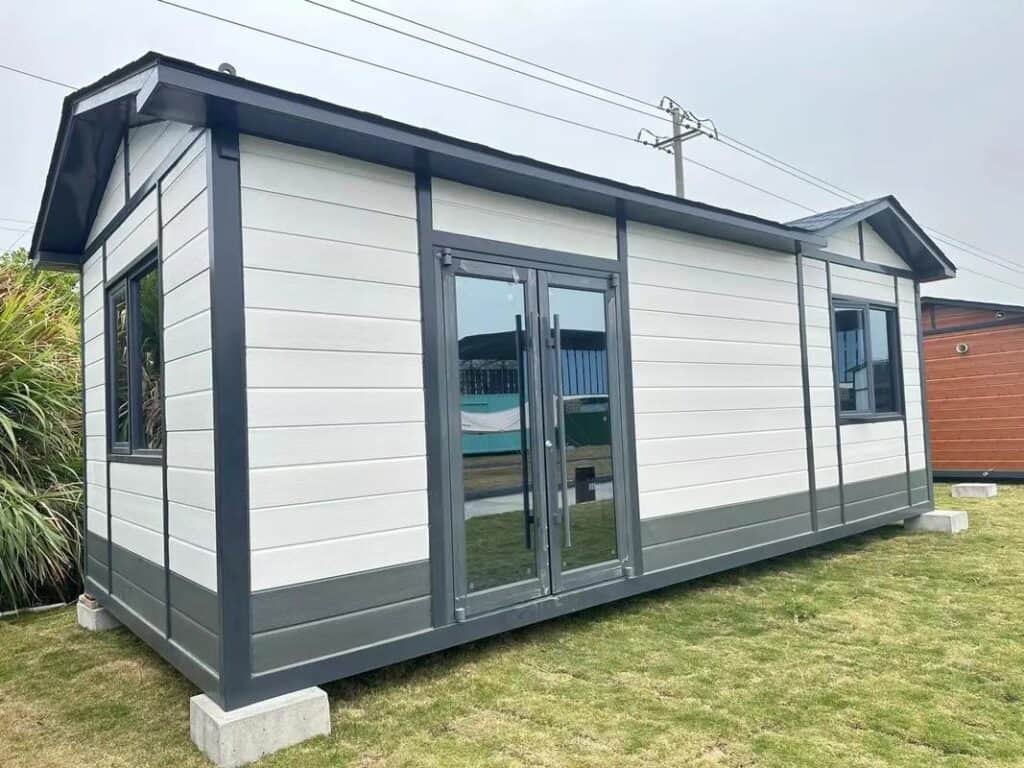 A prefab bungalow with a gable roof, white and gray exterior, large glass doors, and windows, situated on a grassy area with some plants nearby.