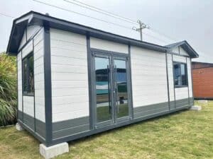 A prefab bungalow with a gable roof, white and gray exterior, large glass doors, and windows, situated on a grassy area with some plants nearby.