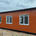 Prefabricated home with a wood-textured exterior and white-framed windows, set in an outdoor environment.