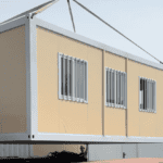 A beige prefab home being unloaded from a truck, with workers in the background for scale.