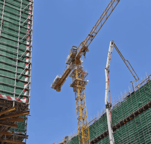 Eine Baustelle mit einem großen gelben Kran, der Materialien hebt. Im Hintergrund ist unter blauem Himmel ein Gebäude mit Gerüst zu sehen.