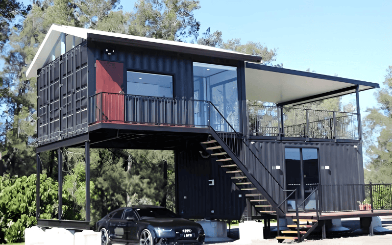 A modern container home elevated on pillars, featuring a staircase leading up to a spacious balcony and a car parked nearby. Surrounded by greenery.