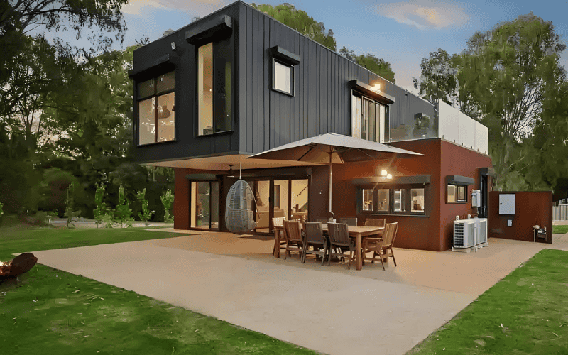 Una elegante casa contenedor con grandes ventanales y balcón, con un fondo de árboles y un cielo azul claro, que ofrece un espacio habitable moderno y compacto.