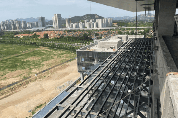 A construction site with steel beams extending outward, overlooking an urban landscape with buildings and greenery.