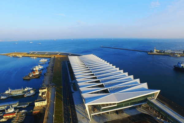 An aerial view of a modern port terminal building with a unique triangular roof design, set beside the sea with ships docked.