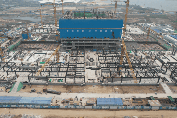 A large industrial construction site featuring a blue central building, steel frames, and multiple cranes in operation.