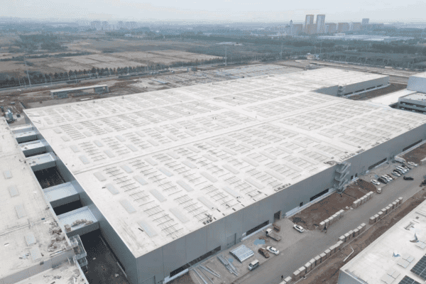 A completed logistics warehouse with a vast white roof, surrounded by roads and greenery.