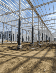 Large steel warehouse under construction, showing exposed beams, columns, and a clear roof structure with natural lighting.