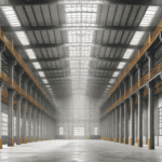 Large industrial warehouse featuring high ceilings, wide open space, and natural light streaming through skylights, supported by strong metal beams.